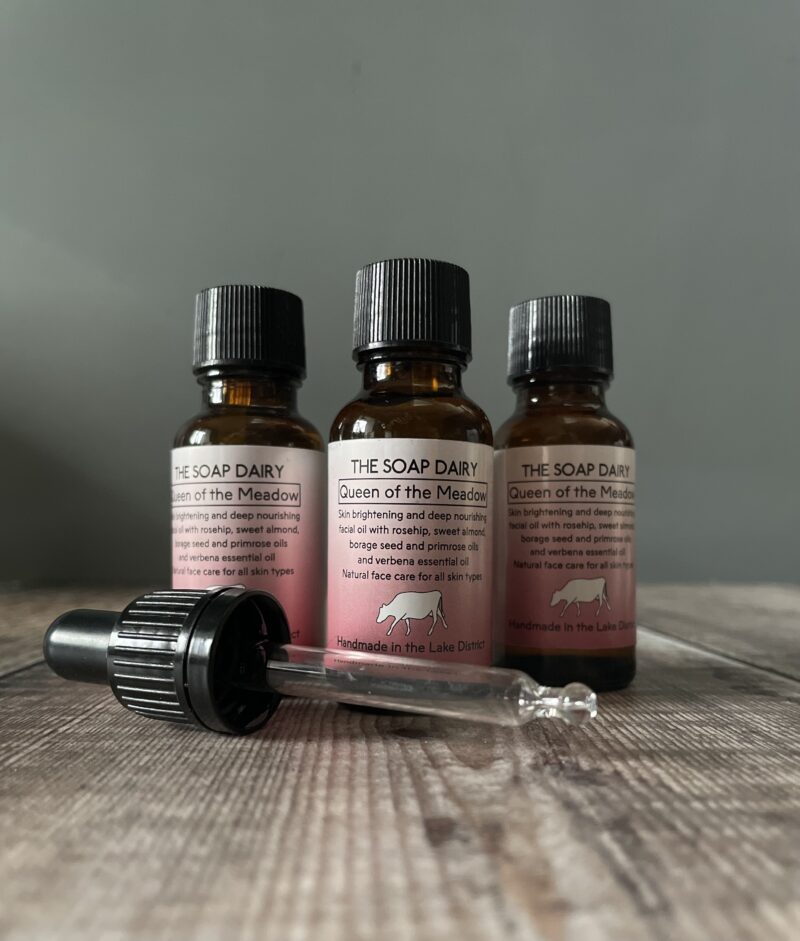Three identical bottles of facial oil with a pink label and a pipette on a wooden board